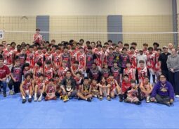 MVP Boys volleyball team and coaches posing together in three rows after a tournament — back row standing, middle row kneeling, front row seated with trophy. Players wear pink, purple, red, and dark MVP jerseys with visible numbers.